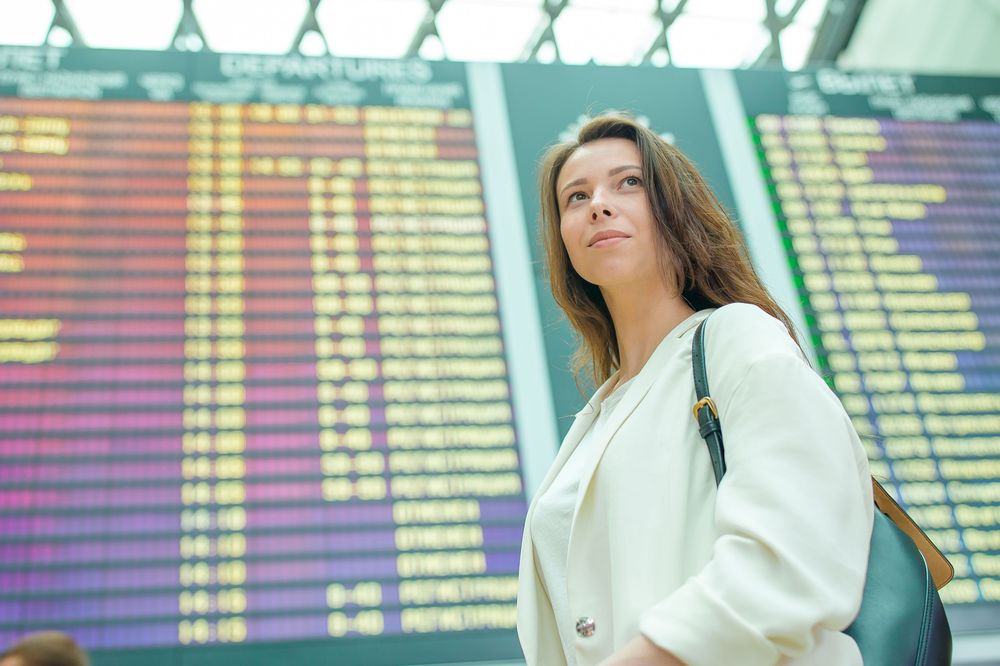  How to save money on flights, young woman on airport in front of flights panel